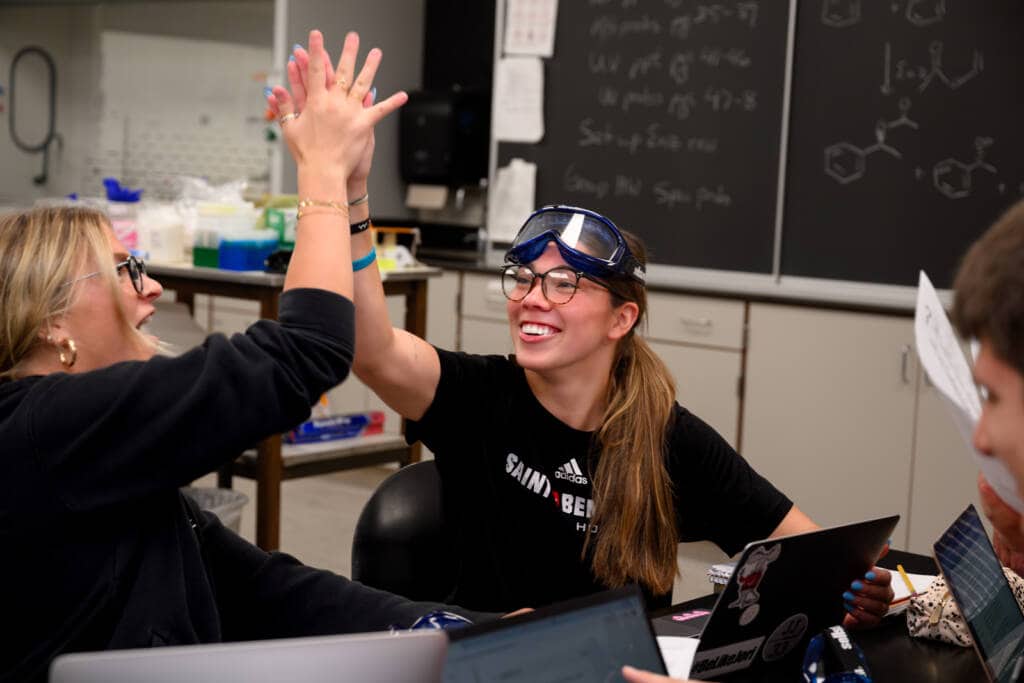 Two students in a chemistry classroom give each other a high-five. One wears safety goggles on her forehead. A chalkboard with chemical formulas is in the background. Laptops and lab equipment are on the table.