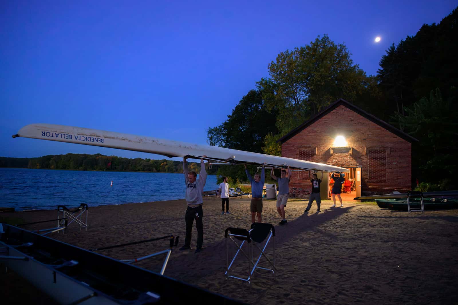 A group of people carries a long rowing boat from a brick boathouse to the edge of a serene lake at dusk. The sky is a deep blue with a visible moon, and the boathouse is warmly lit, illuminating the sandy shore.