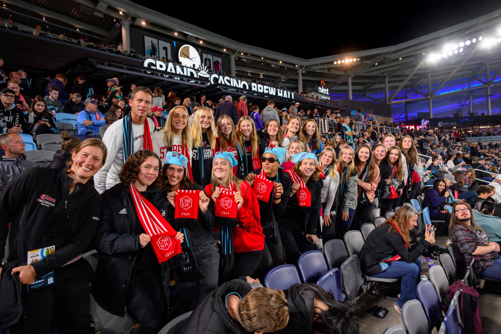 A large group of young people, mostly women, pose for a photo in stadium seats. They are smiling and holding red scarves with a logo. The stadium is well-lit, and other spectators are visible in the background.