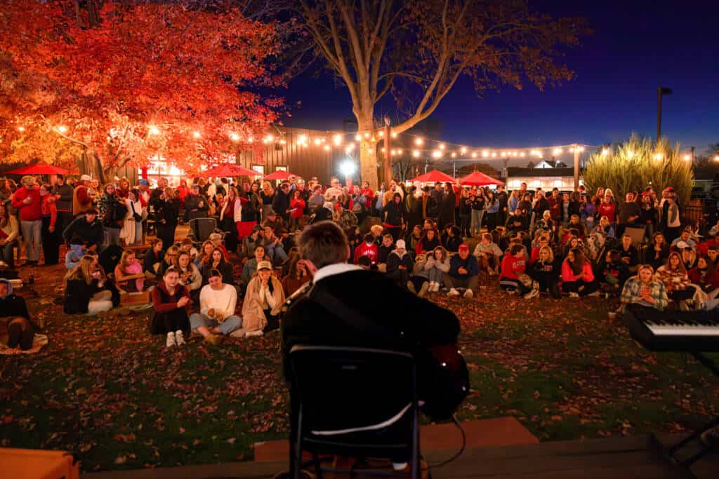 A musician performs on an outdoor stage, facing a large audience seated on the grass. Bright string lights and autumn trees create a warm atmosphere. The audience is dressed in casual, warm clothing, suggesting a chilly evening.