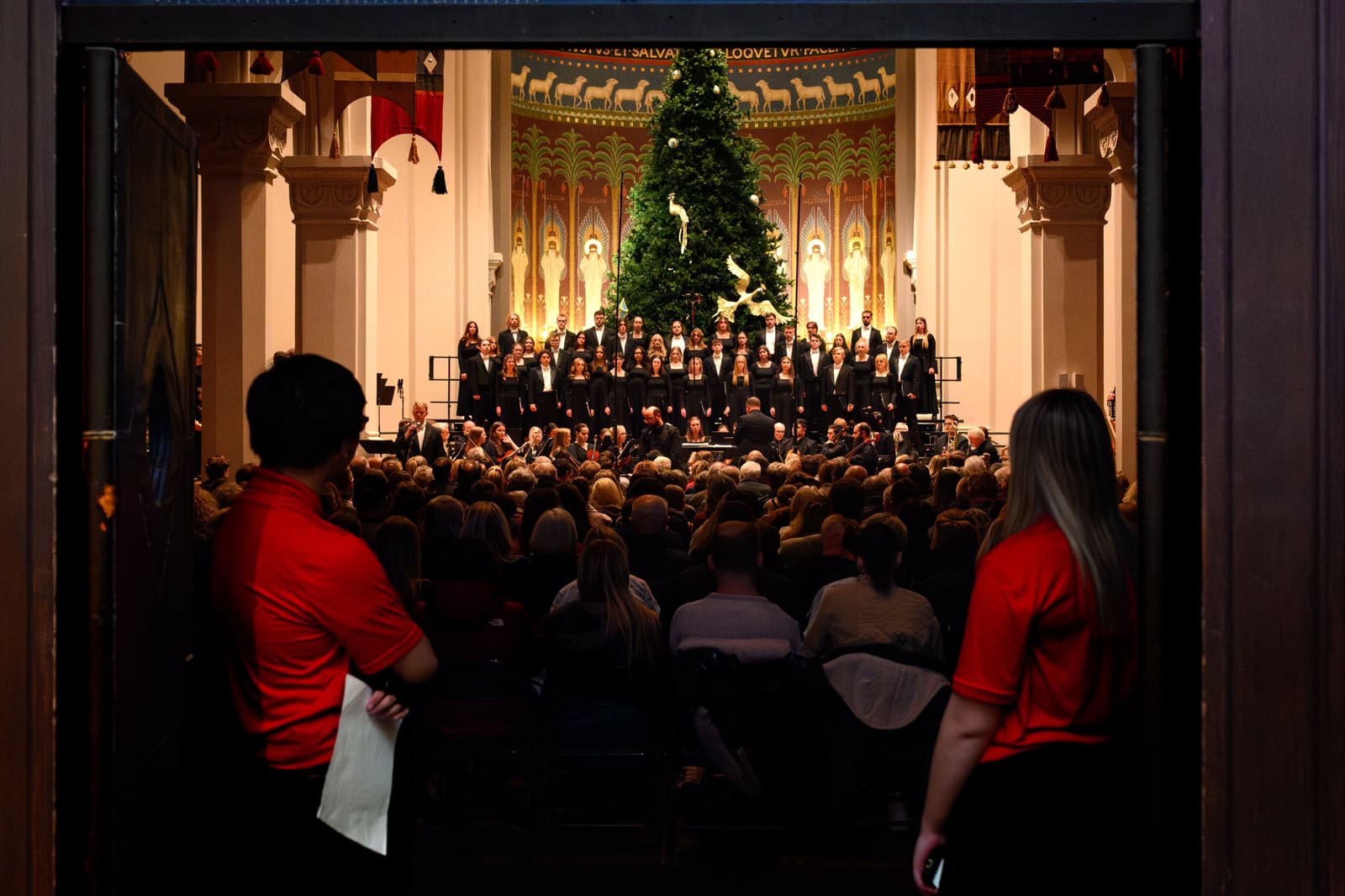A choir dressed in black performs on stage in a large hall decorated with a tall Christmas tree. The audience sits attentively, with two event staff in red shirts standing by the entrance, observing the concert.