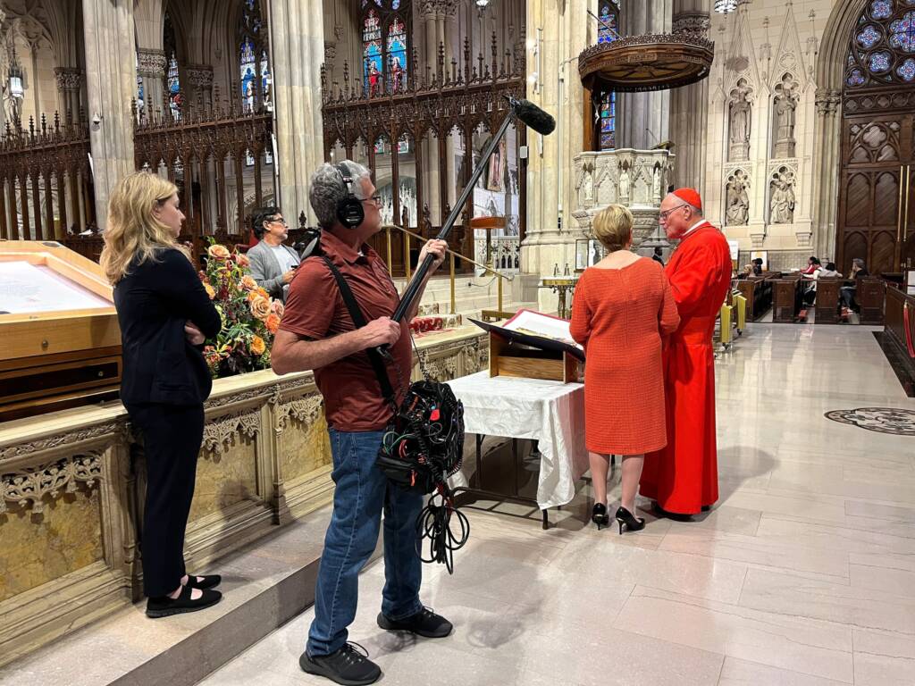 A sound technician holds a boom mic near a cardinal in red robes speaking with a woman in an orange dress inside a cathedral. Two people stand nearby, and ornate architecture with stained glass windows and floral arrangements are visible.