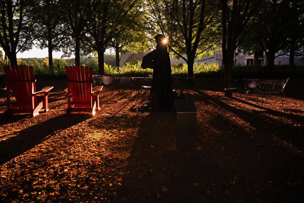 A silhouette of a statue surrounded by red chairs is illuminated by sunlight filtering through trees, casting long shadows on the leaf-covered ground.