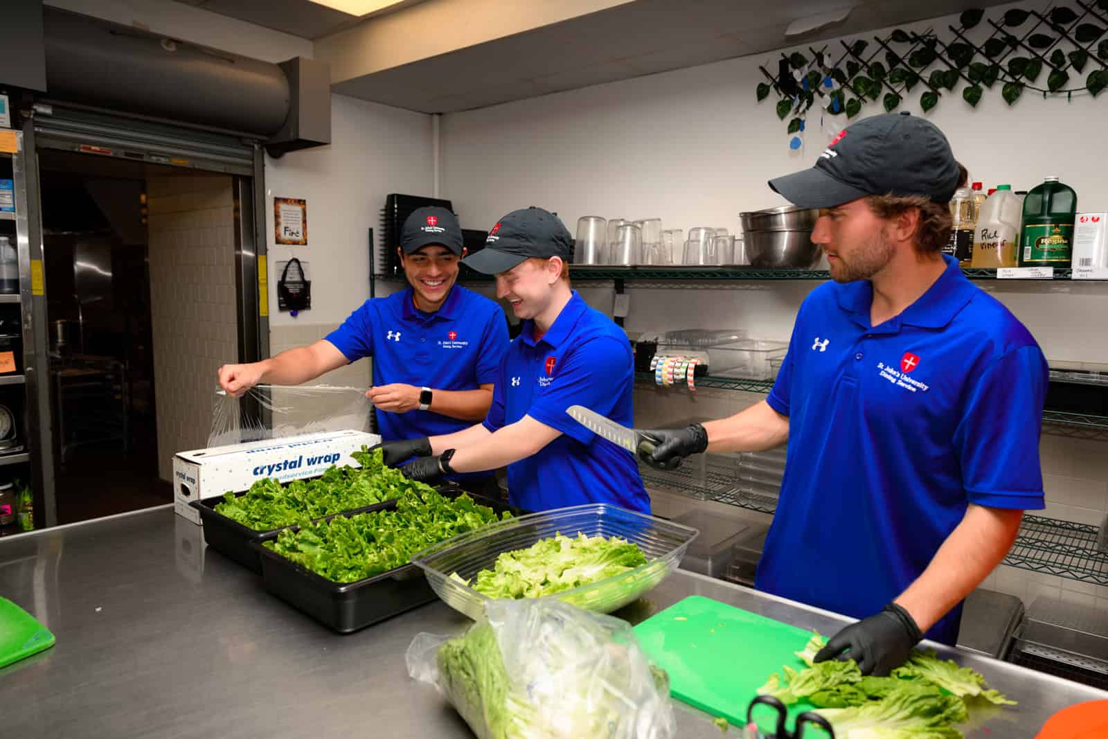 Three people in blue uniforms prepare leafy greens in a commercial kitchen. They are smiling and wearing black gloves as they work, with trays of vegetables on a stainless steel counter.