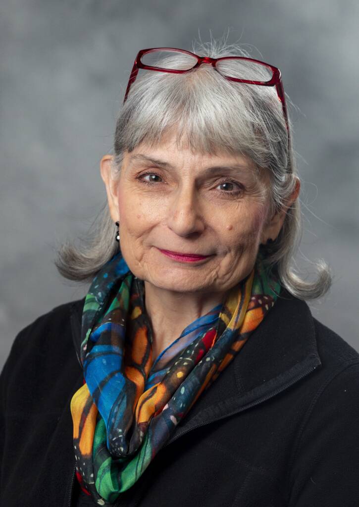 An older woman with gray hair and glasses on her head smiles slightly. She is wearing a colorful scarf and a black top. The background is a muted gray.