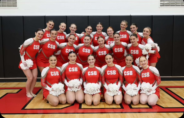 A group of cheerleaders wearing red and white uniforms with "CSB" printed on them pose together in a gym. They are all holding white pom-poms and smiling at the camera, standing on a wood floor. A black mat is visible in the background.