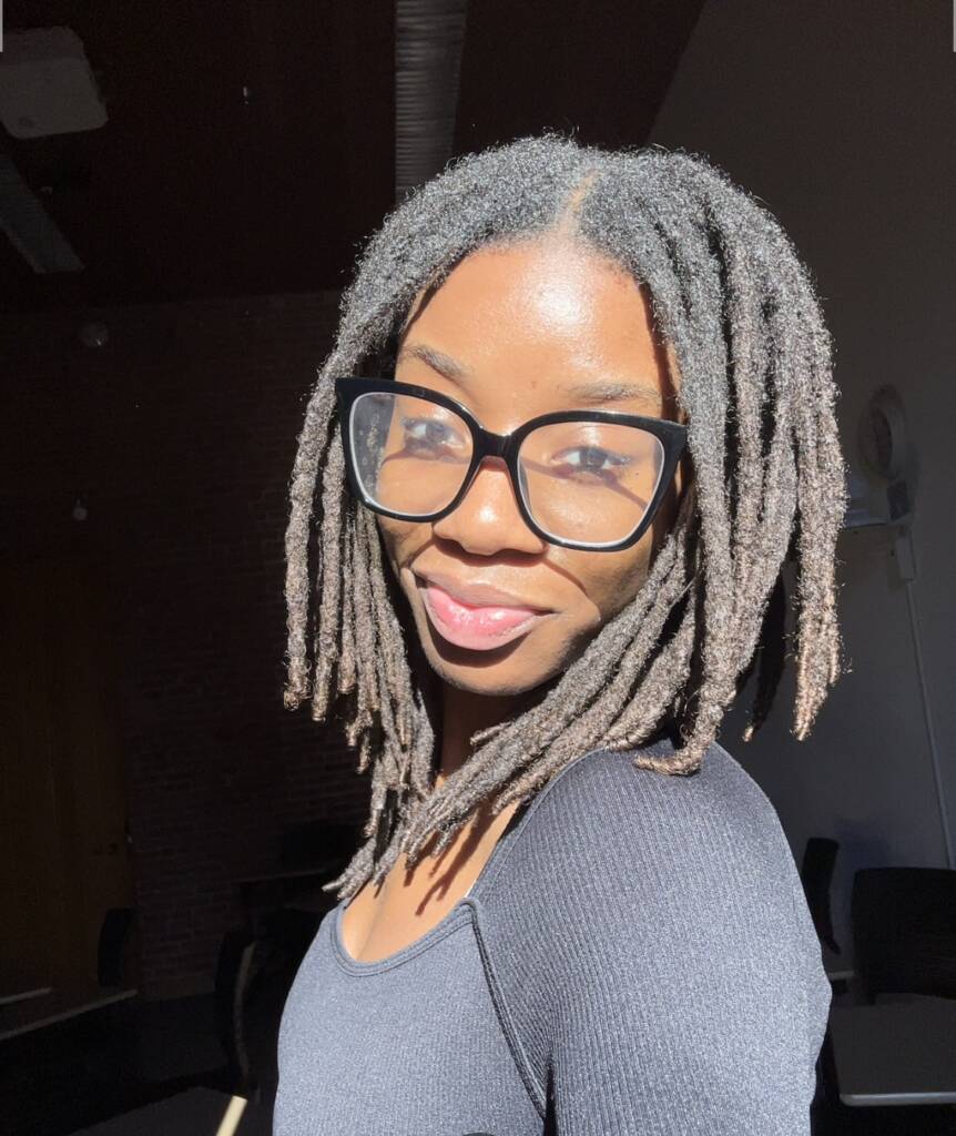 A person with black braided hair and large glasses smiles at the camera. They are wearing a black top and are illuminated by sunlight, standing in a room with a high ceiling and brick wall.