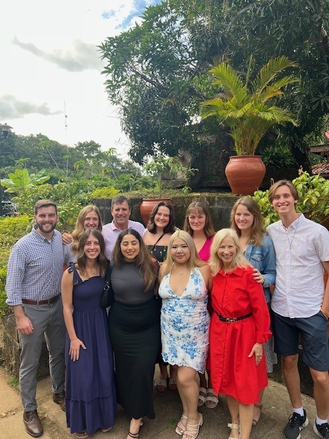 A group of thirteen people, diverse in gender and attire, pose outdoors. They stand in front of a garden with a large potted plant and a small stone structure. The sky is cloudy, and greenery surrounds them.