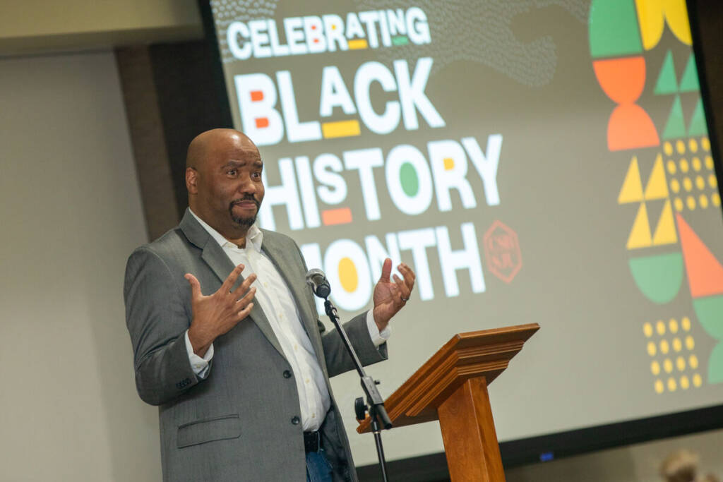 A man in a gray blazer speaks at a podium with a microphone. Behind him, a colorful display reads "Celebrating Black History Month" with abstract patterns.