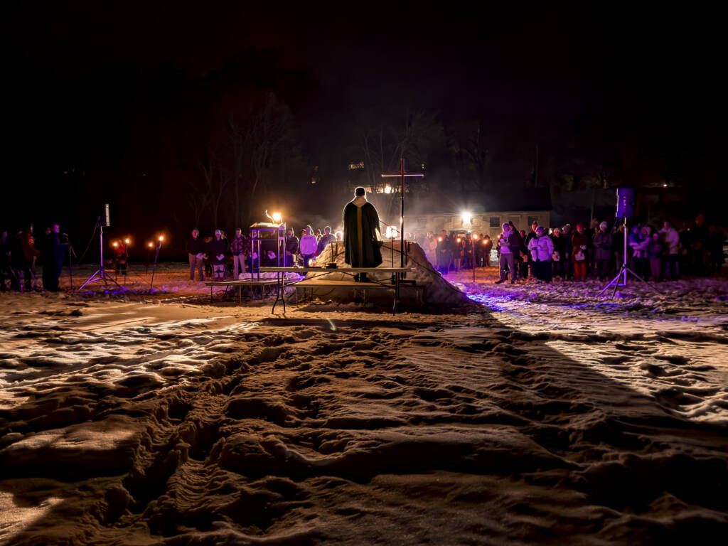 A person on a platform addresses a crowd at night. The ground is snowy, and there are torches flanking the scene. The crowd is gathered in the background, illuminated by the torchlight and a subtle purple glow.