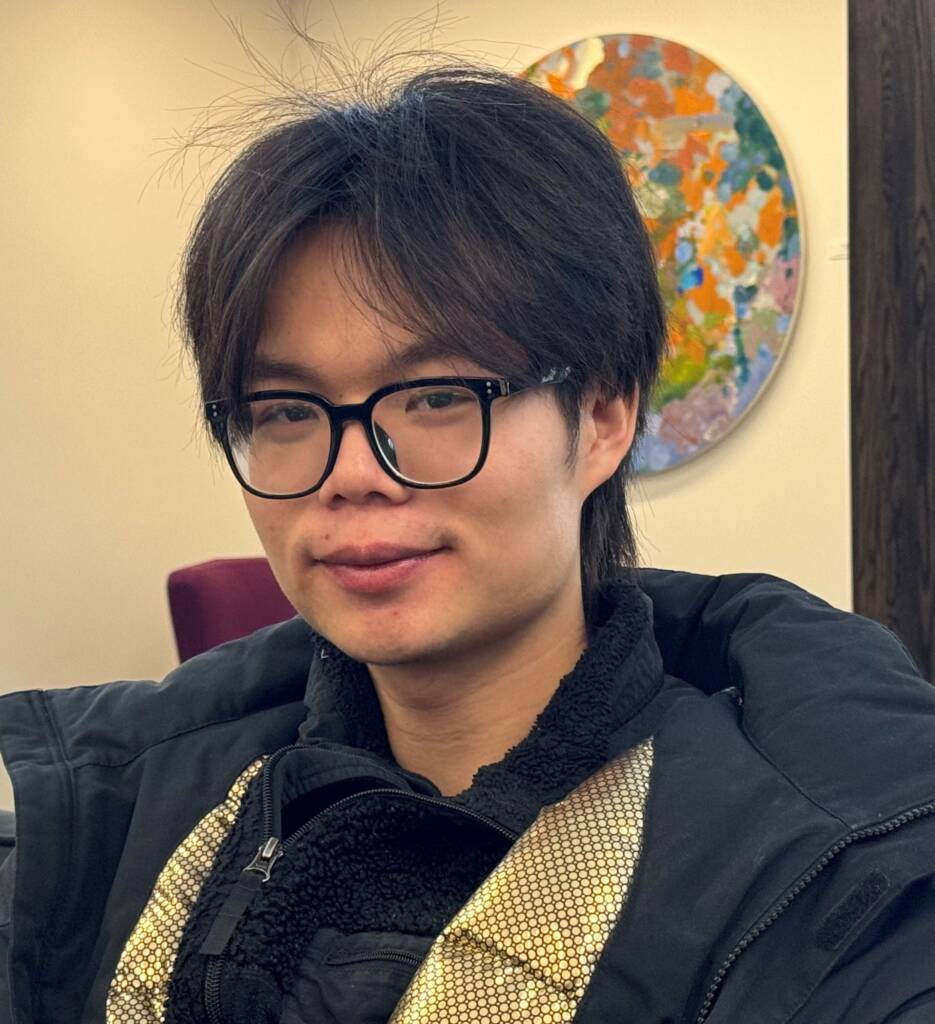 A person with glasses and dark hair is wearing a black jacket with a shiny silver scarf. They are sitting indoors in front of a wall with a colorful, circular abstract artwork.