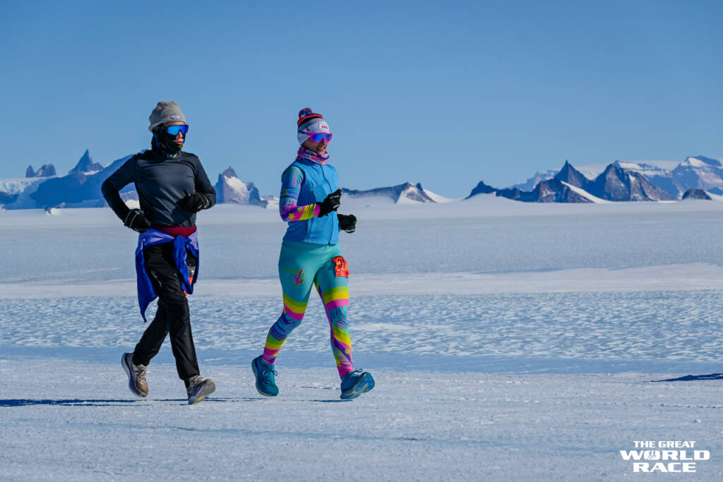 Two people jogging on a snowy landscape with mountainous peaks in the background. They are dressed in colorful winter gear, with hats and sunglasses. The sky is clear and blue. In the corner, there's a logo for "The Great World Race.