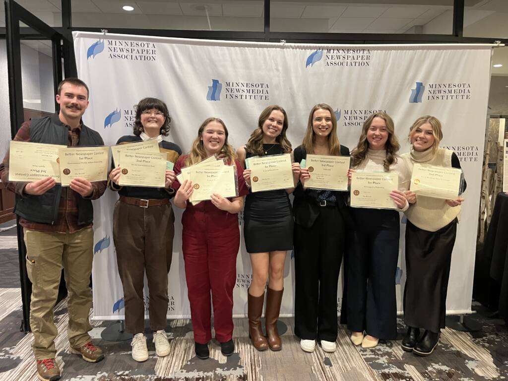Seven people stand side by side, smiling and holding certificates. They are in front of a backdrop with the logos of the Minnesota Newspaper Association and Minnesota News Media Institute.