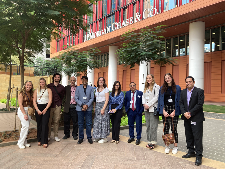 A diverse group of people standing in front of a building labeled "J.P. Morgan Chase & Co." They are outdoors, surrounded by greenery, and wearing business attire with name badges. The group is smiling and posing for the photo.