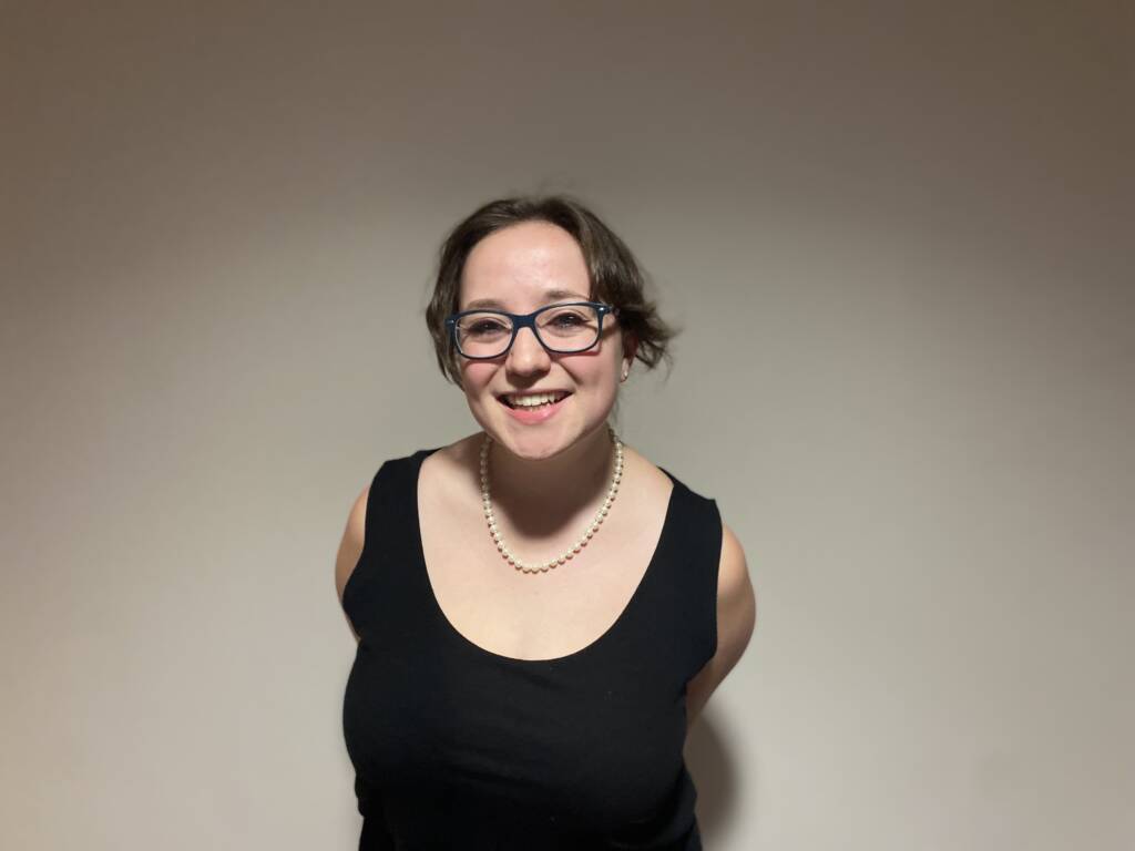 Smiling person with short hair and glasses, wearing a black sleeveless top and a pearl necklace, standing against a plain background.