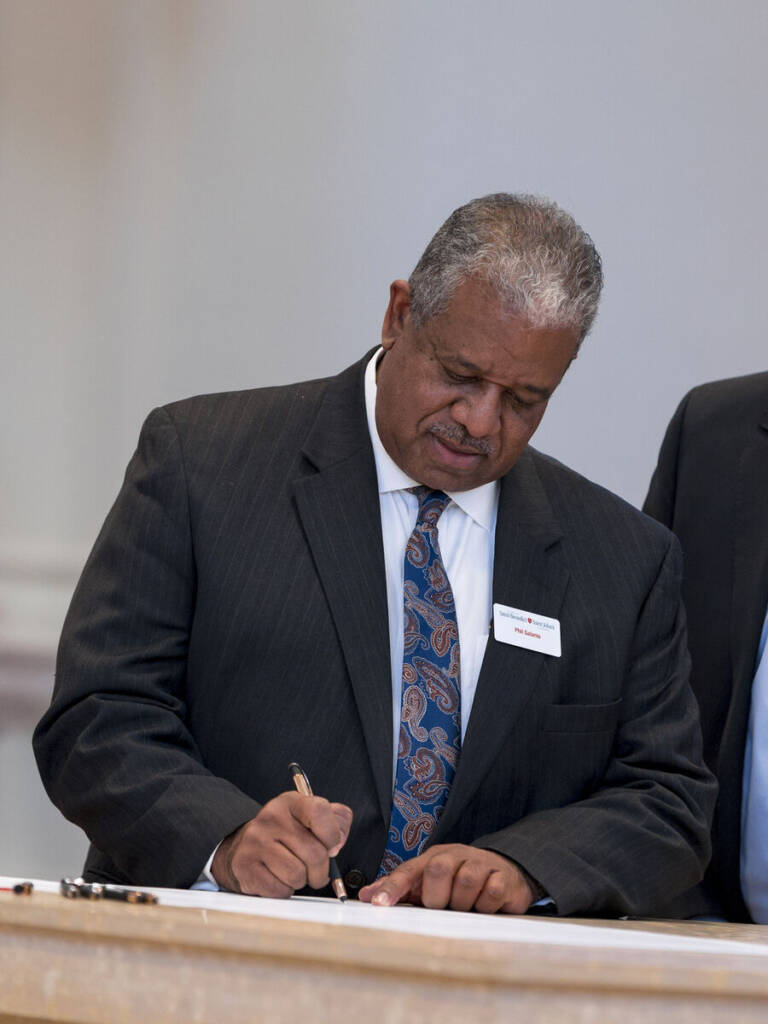 A man in a dark pinstripe suit and paisley tie is standing at a table, writing with a pen on a document. He has short gray hair and wears a name tag on his jacket. The background is plain and blurred.