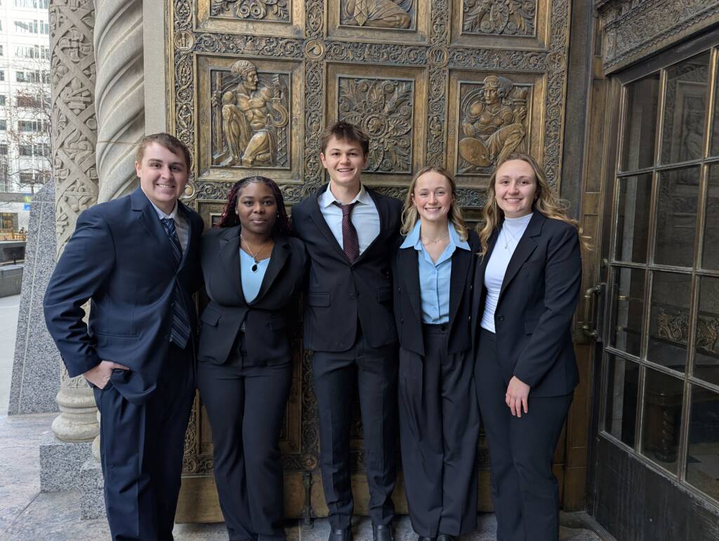 Five people in business attire stand closely together, smiling in front of an ornate, sculpted door. The group includes two men and three women, each wearing a suit. The background shows part of a stone building and intricate door design.