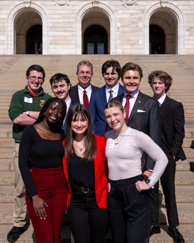 A diverse group of nine young adults, dressed in business casual attire, stand on the steps of a grand building with stone arches. The group is smiling and posing for the photo, suggesting a formal or celebratory occasion.