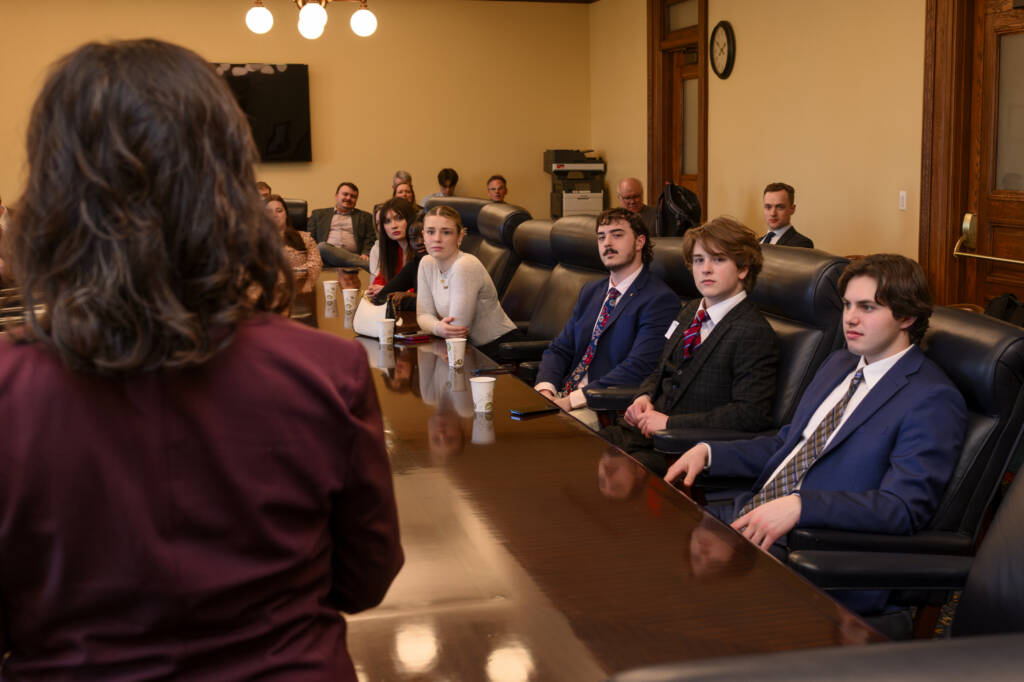 A group of people in formal attire sit around a long table in a conference room. One person stands at the head of the table, addressing the group. A few people in the background are observing.