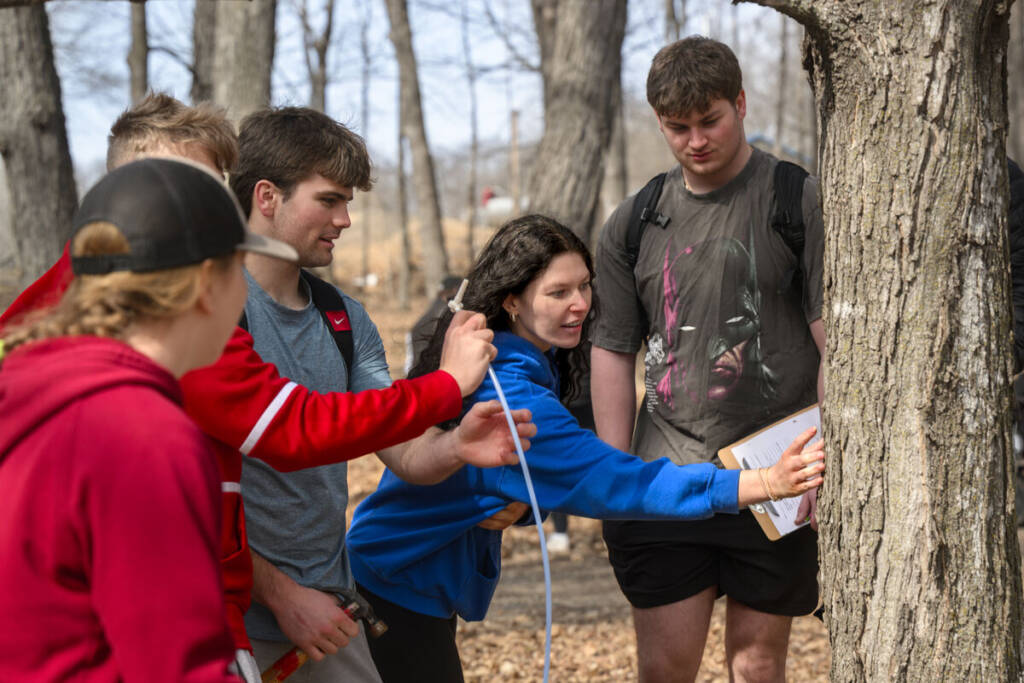 A group of young adults gathers around a tree in a forest. One person points at the tree while another holds a long tube. A person in a red shirt holds a tool. They appear to be engaging in an outdoor educational activity.