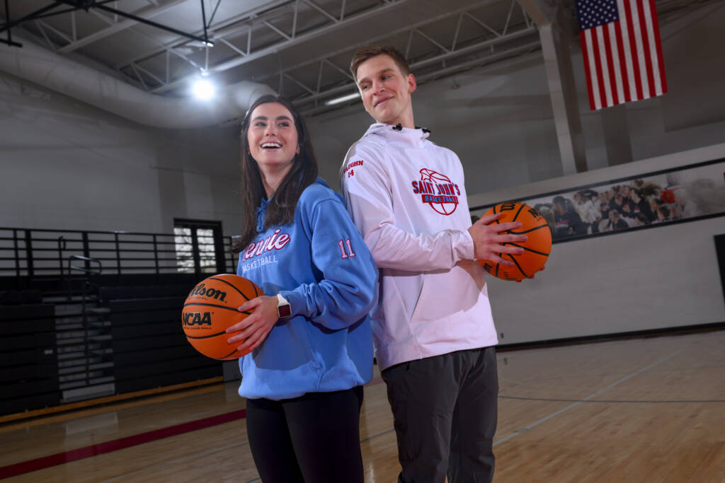 Two people stand back-to-back in a gym, each holding a basketball. One wears a light blue hoodie and the other a white sweater. An American flag hangs in the background, and a mural is on the wall. They smile, looking in opposite directions.