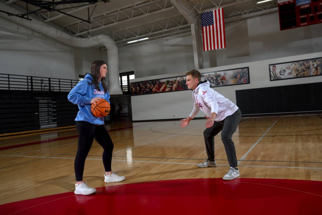 A young woman in a blue hoodie holds a basketball, facing a young man in a white hoodie on a gymnasium court. The court has a red center circle, and a U.S. flag is displayed above. Both appear focused and engaged in a game.