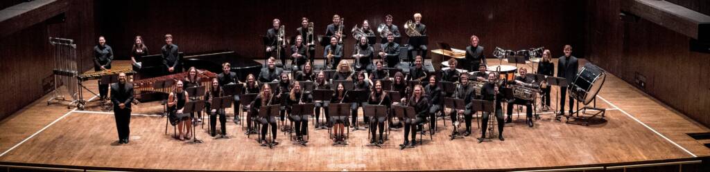 A large orchestra seated on stage, with musicians holding various instruments such as brass, woodwinds, and percussion. They are arranged in neat rows, and the stage is lit with bright overhead lights, framed by dark wooden walls.