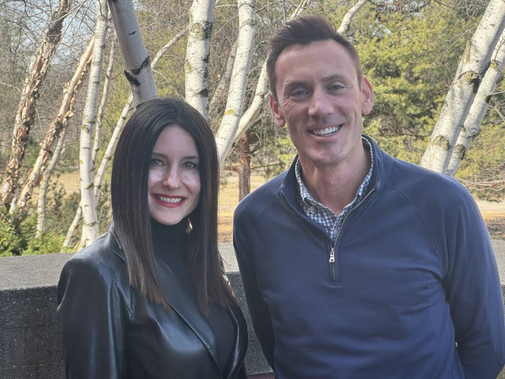 Two people are smiling outdoors with trees in the background. The person on the left has long dark hair and is wearing a black jacket. The person on the right has short hair and is wearing a dark pullover over a checkered shirt.