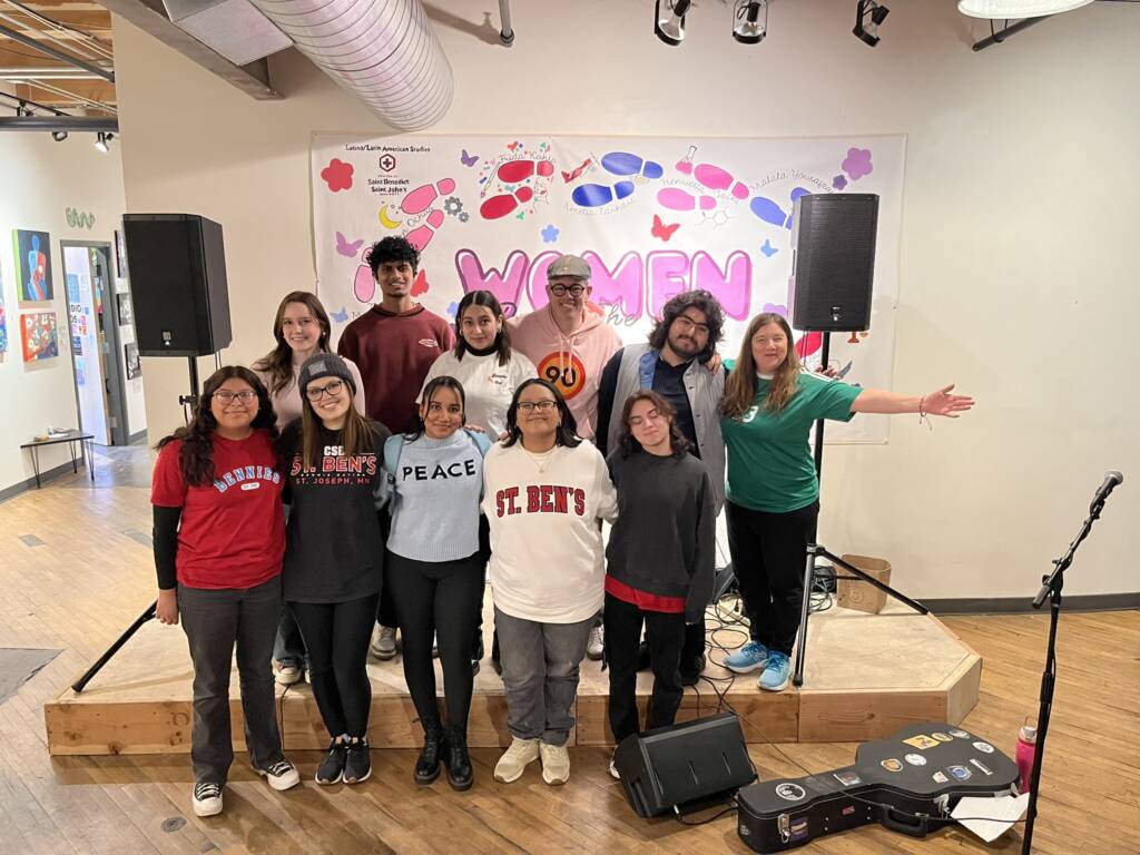 A group of smiling people stand on a wooden stage indoors. They are wearing casual clothes with various logos. Behind them is a banner with "Women" in colorful letters, decorated with flowers and butterflies. Musical equipment is visible around the stage.