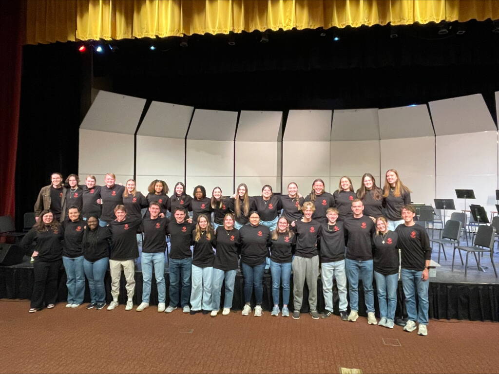 A group of people wearing matching black shirts with a red logo stand together on a stage in a theater. The background shows a curved, partitioned backdrop. The theater has red curtains and a yellow valance above the stage.