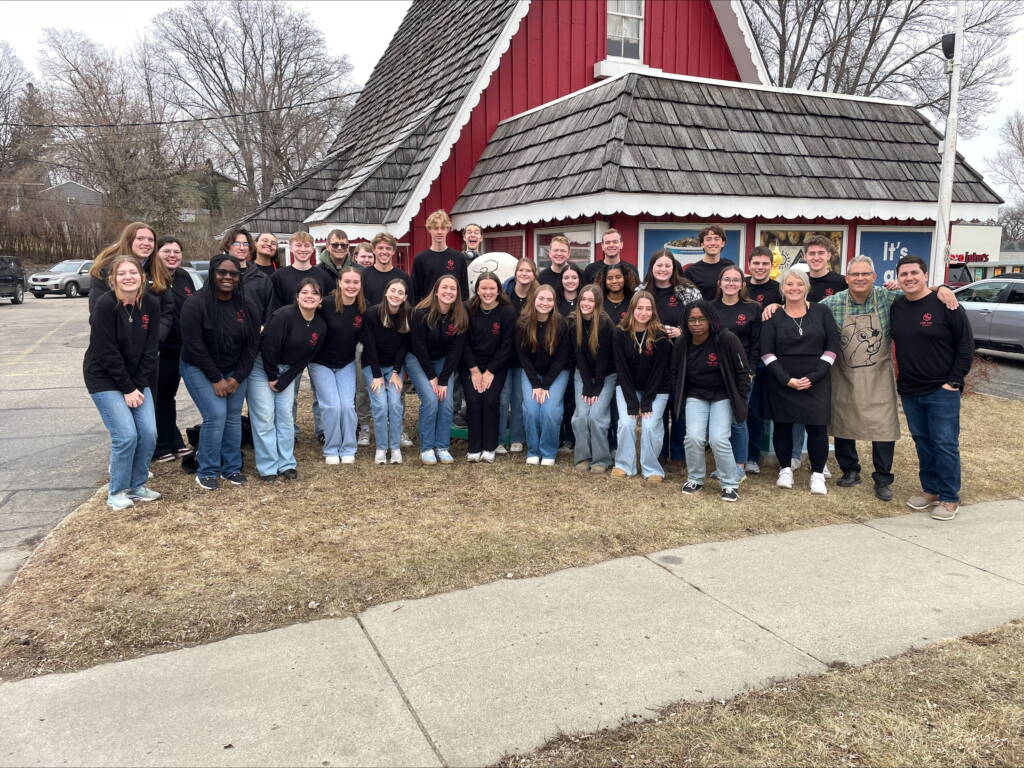 A large group of people wearing black shirts and jeans stand together, smiling, in front of a red building with a shingled roof. Two people in aprons stand among them. Trees and a cloudy sky are in the background.
