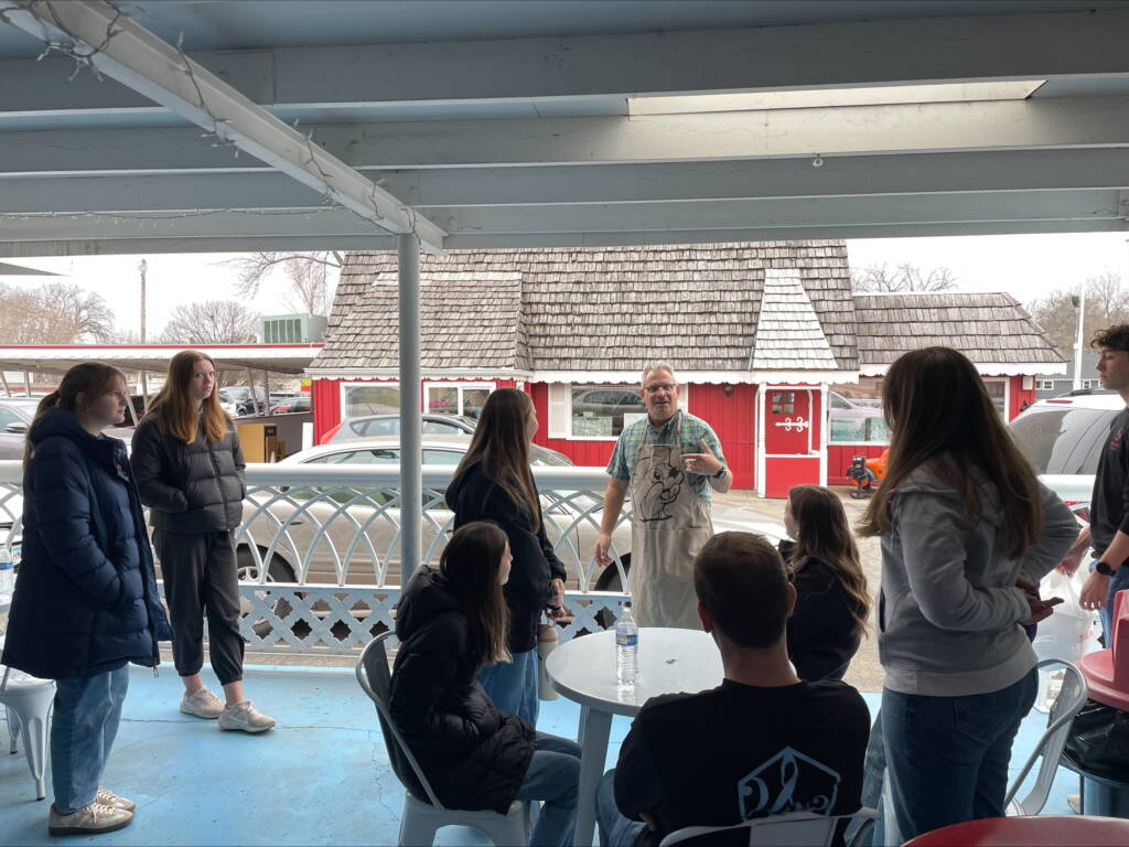 A group of people gather on a covered porch. A person wearing an apron stands and speaks to them. Red buildings with shingled roofs are visible in the background. The group listens attentively, seated and standing around tables.