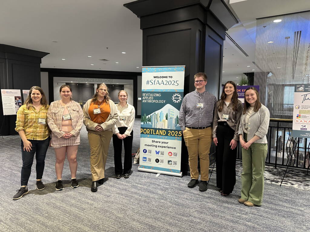 A group of people standing indoors beside a banner that reads, "Welcome to SFAA 2025: Revitalizing Applied Anthropology, Portland 2025." They are smiling and dressed in casual to business casual attire. The background shows a spacious interior setting.