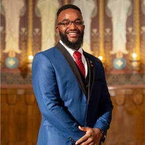 A man in a blue suit with a red tie smiles while standing in an ornate room with candles and decorative wall details.