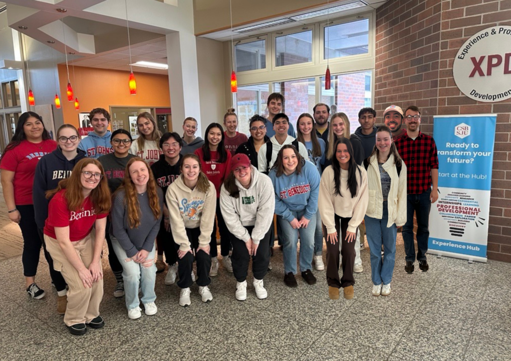 A group of students stand together smiling indoors near a sign that reads "Ready to transform your future? Start at the Hub," in a brightly lit hallway with red pendant lights and XPDerience Hub signage.