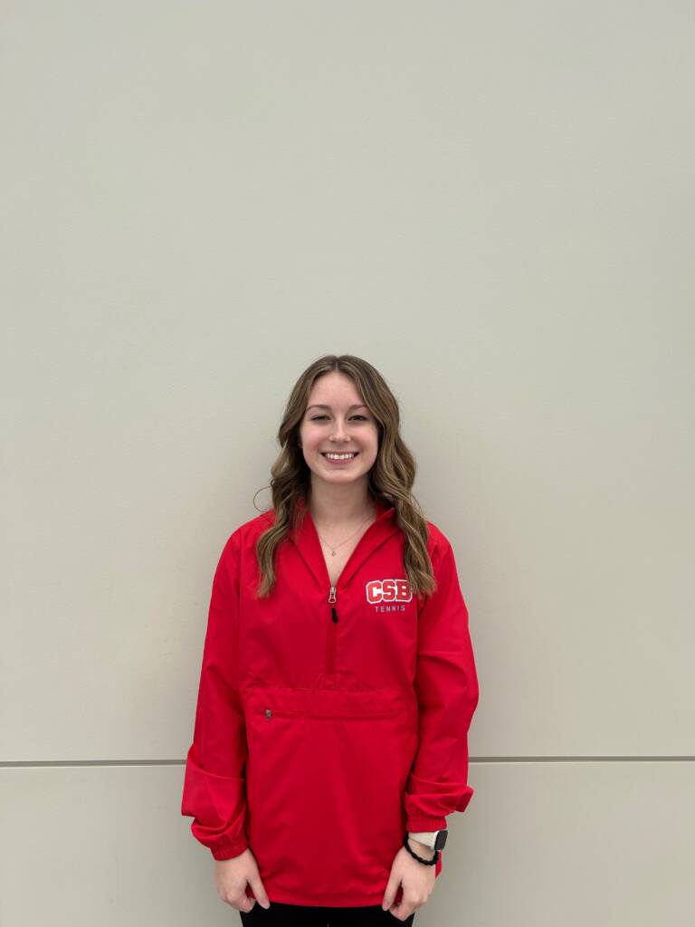 A young woman with long wavy hair stands smiling against a plain light-colored wall, wearing a bright red jacket with "CSB Tennis" printed on it.