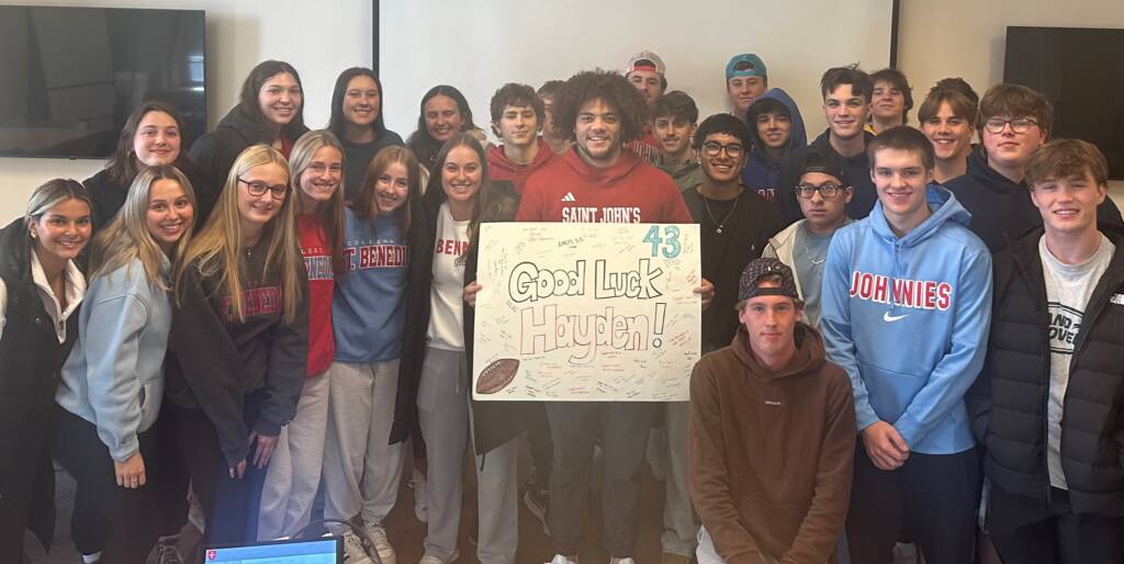 A group of students poses indoors, smiling around a large handmade sign that reads “Good Luck Hayden!” with a football drawing and the number 43. Two large TVs are on the wall in the background.