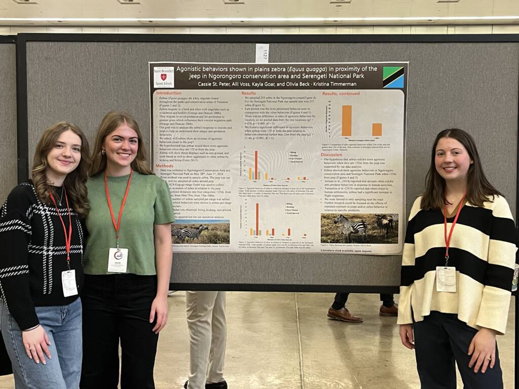Three young women stand smiling in front of a scientific poster about zebra behavior, displayed on a gray board at a conference. The poster features text, graphs, and photos related to their research.