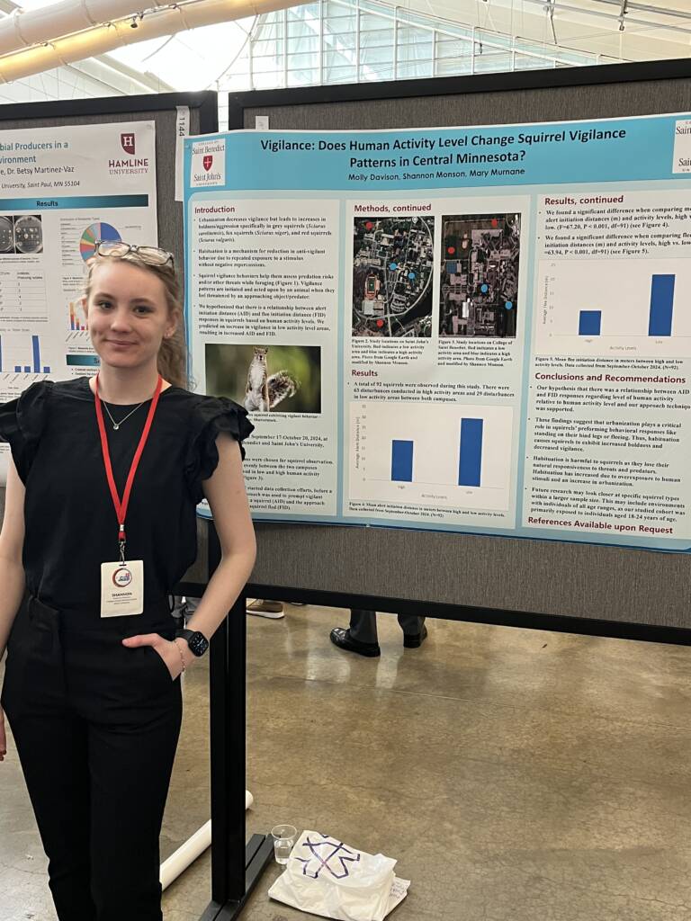A young woman stands beside a scientific poster titled "Vigilance: Does Human Activity Level Change Squirrel Vigilance Patterns in Central Minnesota?" in an indoor event space. She is smiling and wearing a badge.