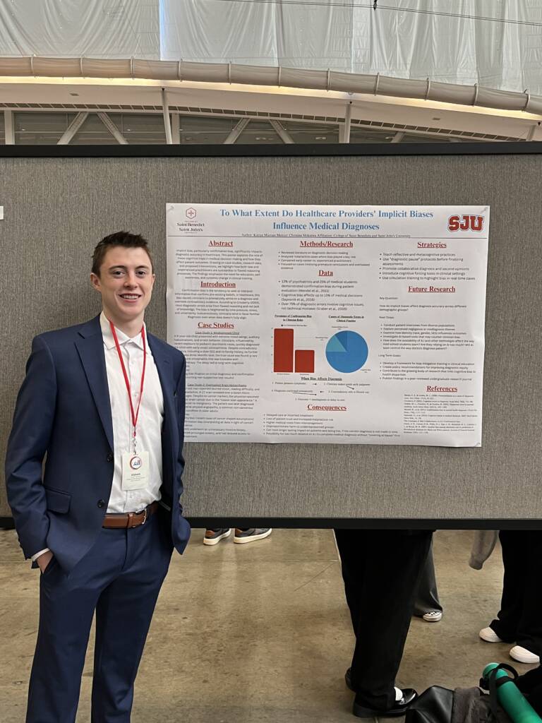 A young man in a suit stands smiling next to a scientific research poster titled "To What Extent Do Healthcare Providers' Implicit Biases Influence Medical Diagnoses" at an academic event.