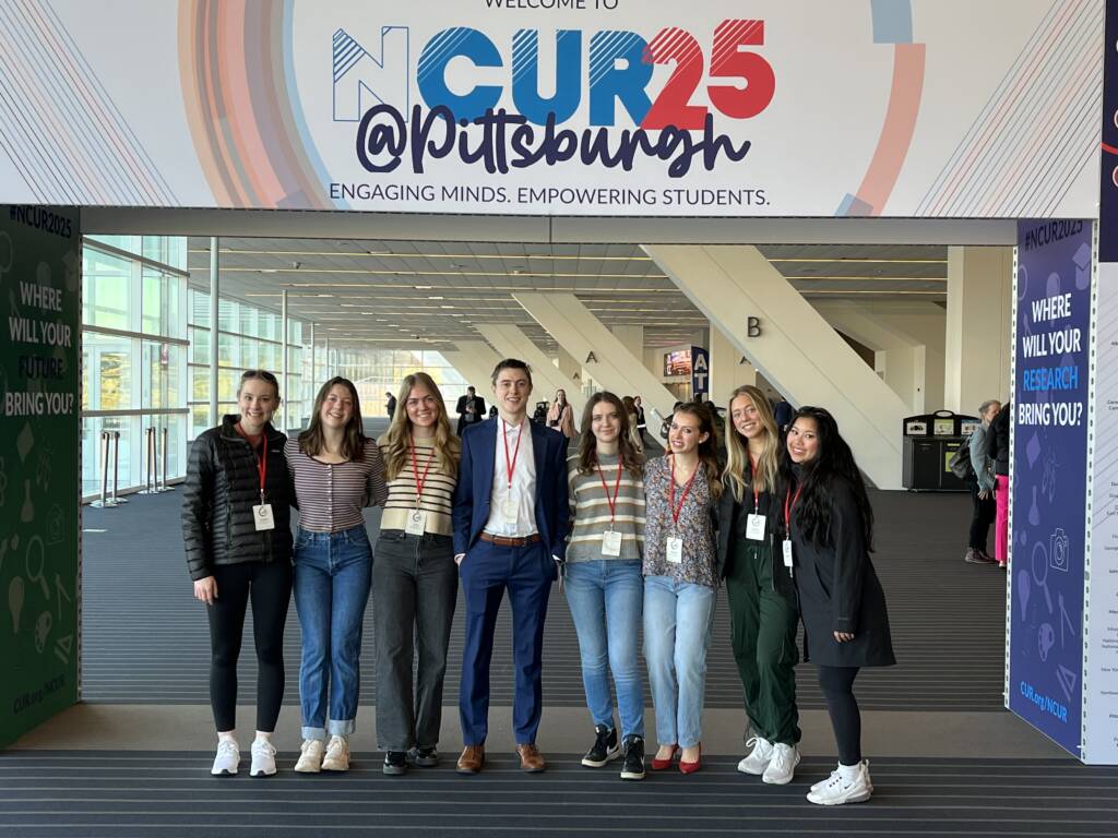 A group of nine young adults stand smiling in a row under a large sign reading "CUR 25 @ Pittsburgh" in a bright convention center hallway. Most are casually dressed, with some wearing name tags and lanyards.