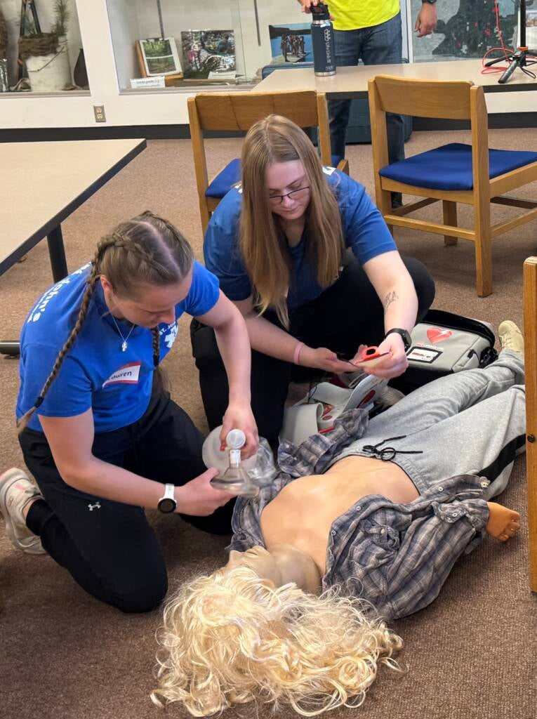 Two people in blue shirts practice CPR on a training mannequin. One person uses a bag valve mask, while the other prepares an automated external defibrillator (AED). Chairs and display cases are visible in the background.