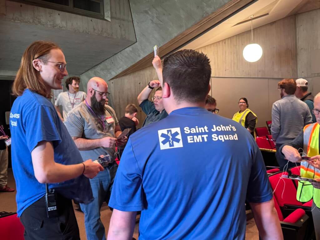 A group of people is gathered in a room with red chairs and concrete walls. Two individuals in blue shirts with "Saint John's EMT Squad" written on the back are in the foreground. One person holds a card up in the air.