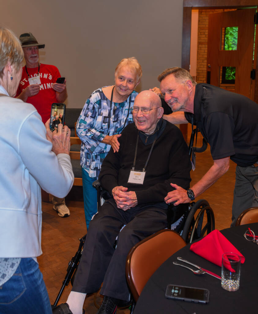 A group of people smiling as a person in a wheelchair poses for a photo. A woman is taking the picture with a phone. They are indoors near a table. Everyone appears to be having a joyful moment together.
