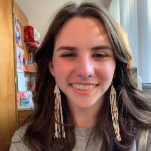 A person with long brown hair smiles in a room. They wear long, beaded earrings and a nose ring. Sunlight illuminates their face. The background includes wooden furniture, wall decorations, and a partially open window.