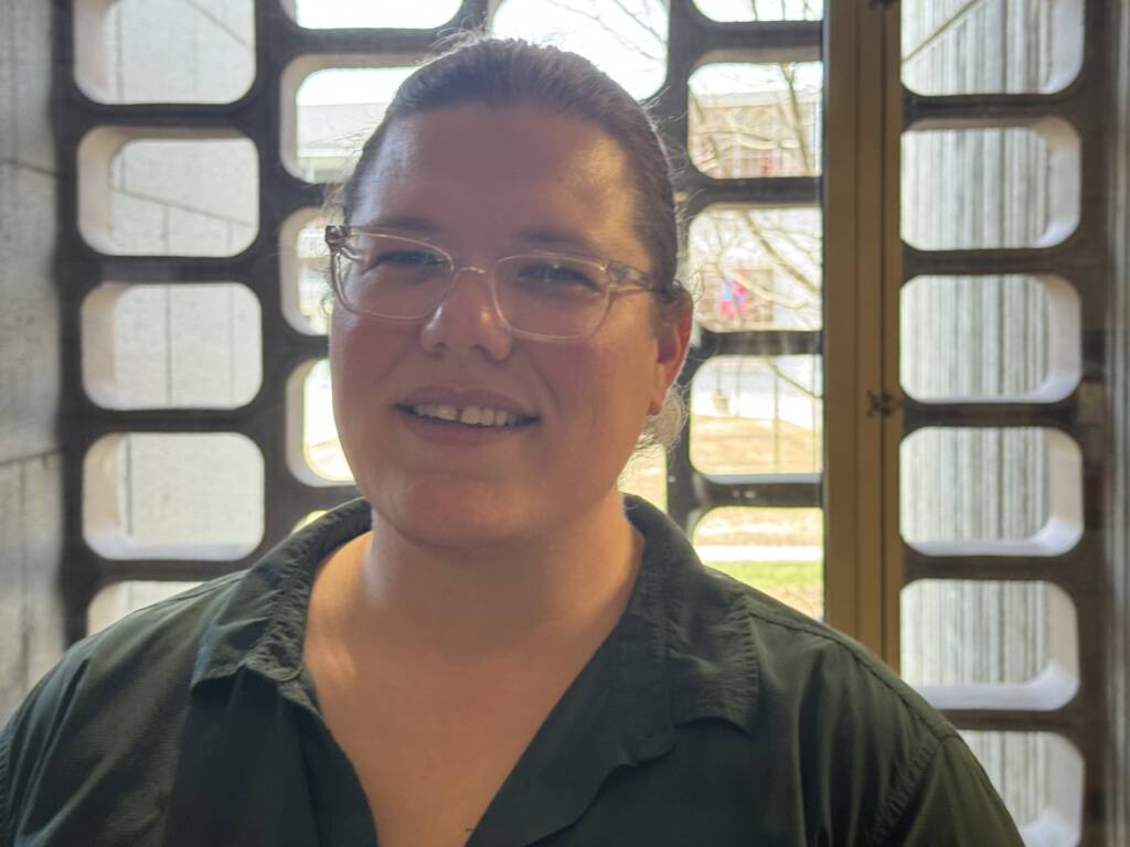 A person with glasses and long hair tied back is smiling indoors. They're wearing a green shirt and standing in front of a window with a decorative grille, allowing diffused light to come through. The background appears to show a building exterior.