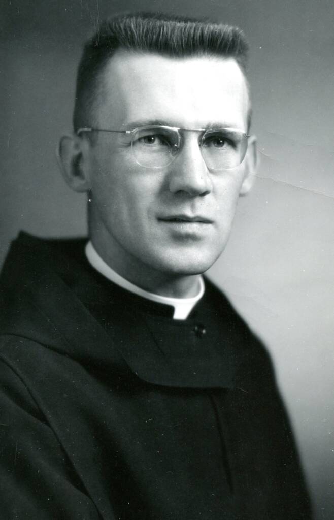 Black and white portrait of a man wearing glasses and a clerical collar. He has short hair and is dressed in religious attire, looking straight at the camera with a neutral expression.
