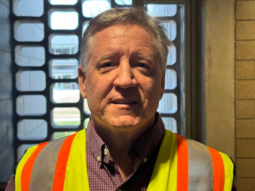 A person wearing a safety vest stands indoors near a window with a grid pattern. They are dressed in a checkered shirt and smiling slightly at the camera. The lighting creates a shadowed effect on the person's face.