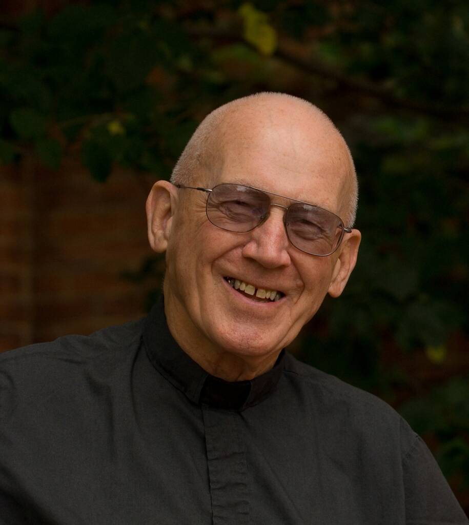 Smiling older man with glasses and a clerical collar, wearing a dark shirt. He is outdoors with a blurred background of green leaves and a brick wall.