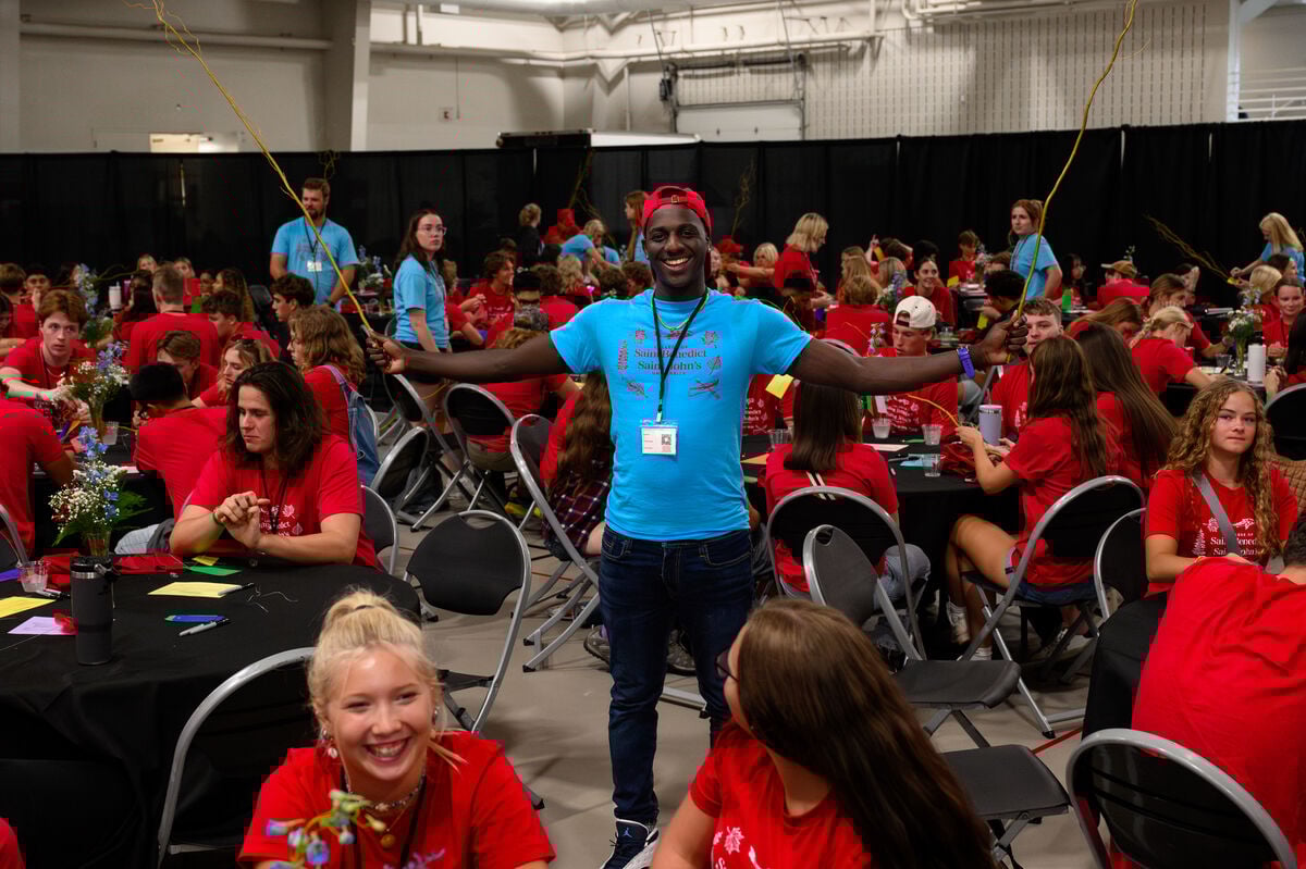 A smiling man in a blue shirt and red cap stands with arms outstretched, holding balloons, surrounded by people in red shirts seated at round tables in a busy indoor event space.